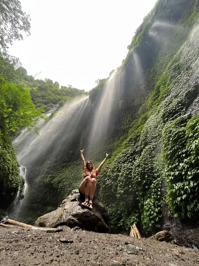 Una mujer sentada en una roca con los brazos en alto, frente a una cascada que desciende por un acantilado verde y exuberante.