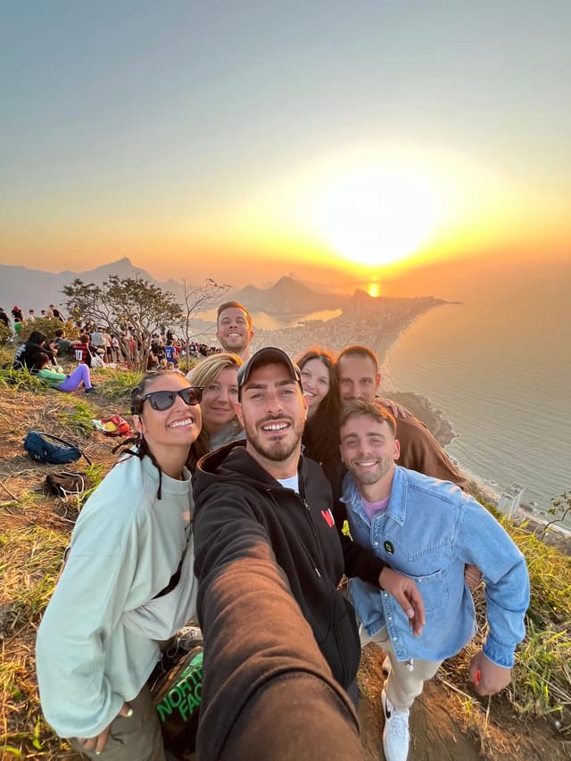 Un voyage en groupe WeRoad prend un selfie souriant depuis un point de vue dominant une ville côtière et des montagnes au coucher du soleil.