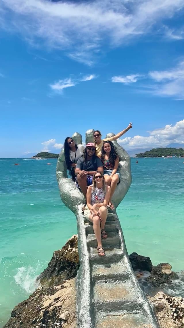 A WeRoad group trip of people posing for a photo on a large, hand-shaped sculpture next to the turquoise sea.