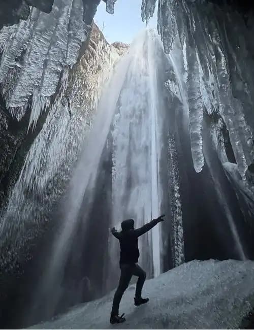 A person with arms outstretched stands on an icy slope at the base of a large, partially frozen waterfall surrounded by icicles.