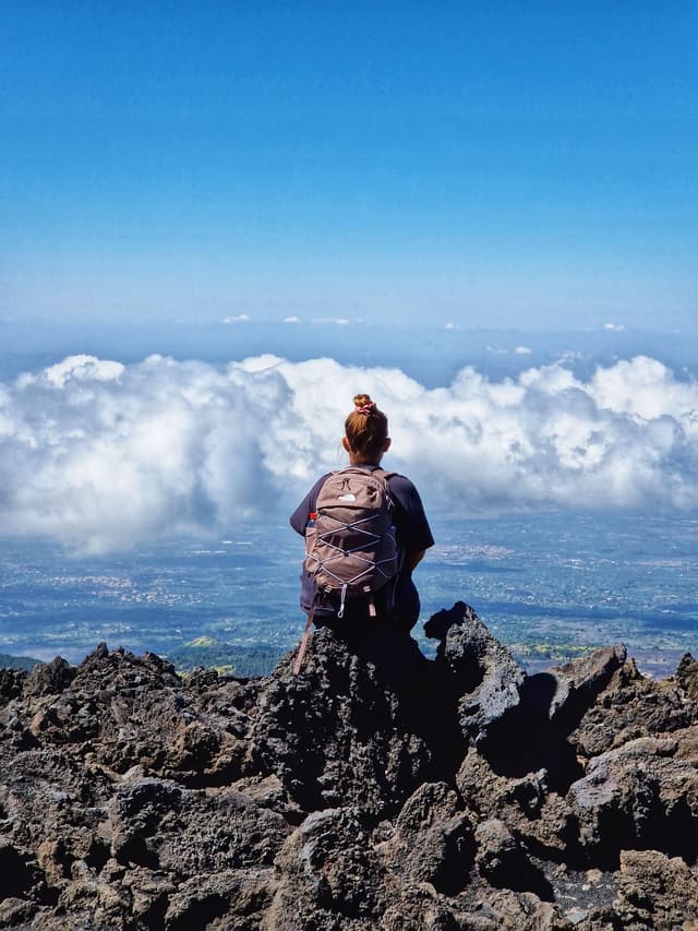 Eine Person mit Rucksack sitzt auf einem zerklüfteten Vulkanfelsen und blickt von einem Berggipfel auf ein Wolkenmeer.