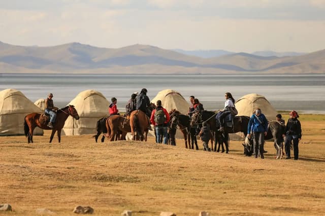 Un grupo de viajeros a caballo junto a un campamento de yurtas a orillas de un lago entre montañas.