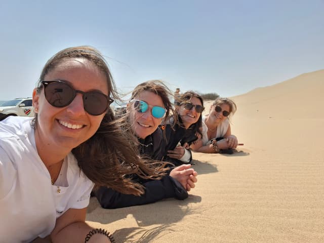 Un voyage de groupe WeRoad de quatre femmes portant des lunettes de soleil allongées sur une dune de sable sous un ciel ensoleillé.
