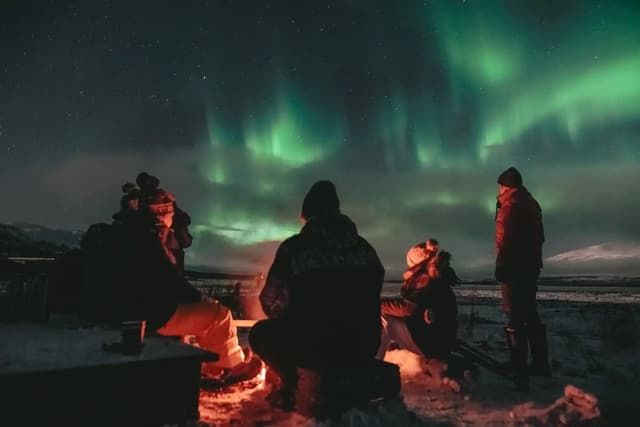 Viajeros junto a una hoguera bajo la aurora boreal en una noche nevada.