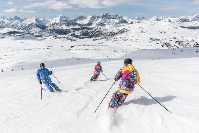 Eine WeRoad-Gruppenreise von Skifahrern in bunten Anzügen, die einen schneebedeckten Hang mit Blick auf riesige Gipfel hinabfahren