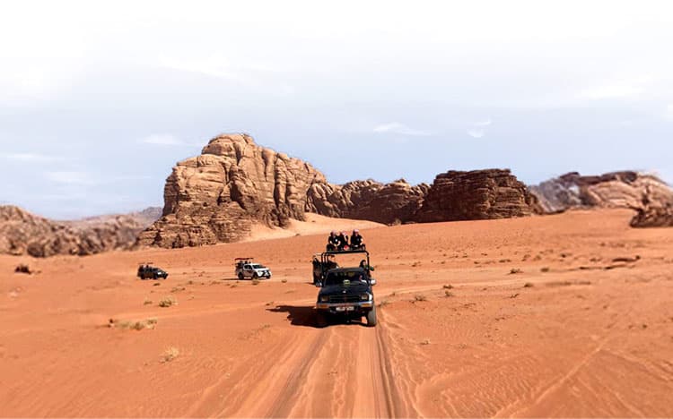 Convoglio di fuoristrada attraversa la sabbia rossa del deserto tra formazioni rocciose e cielo limpido.